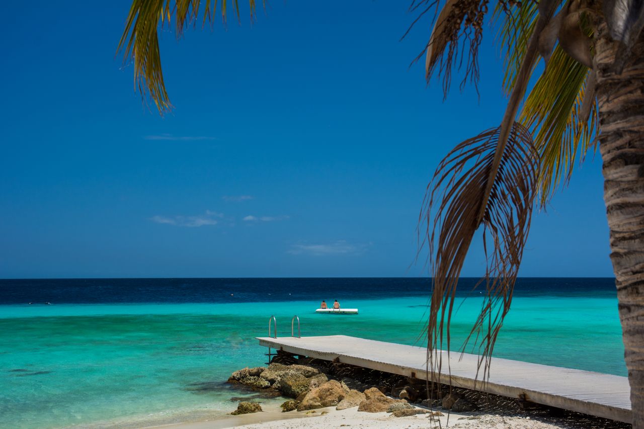 portfolio-04 A tranquil beach scene with a wooden pier extending into turquoise water under a clear blue sky.